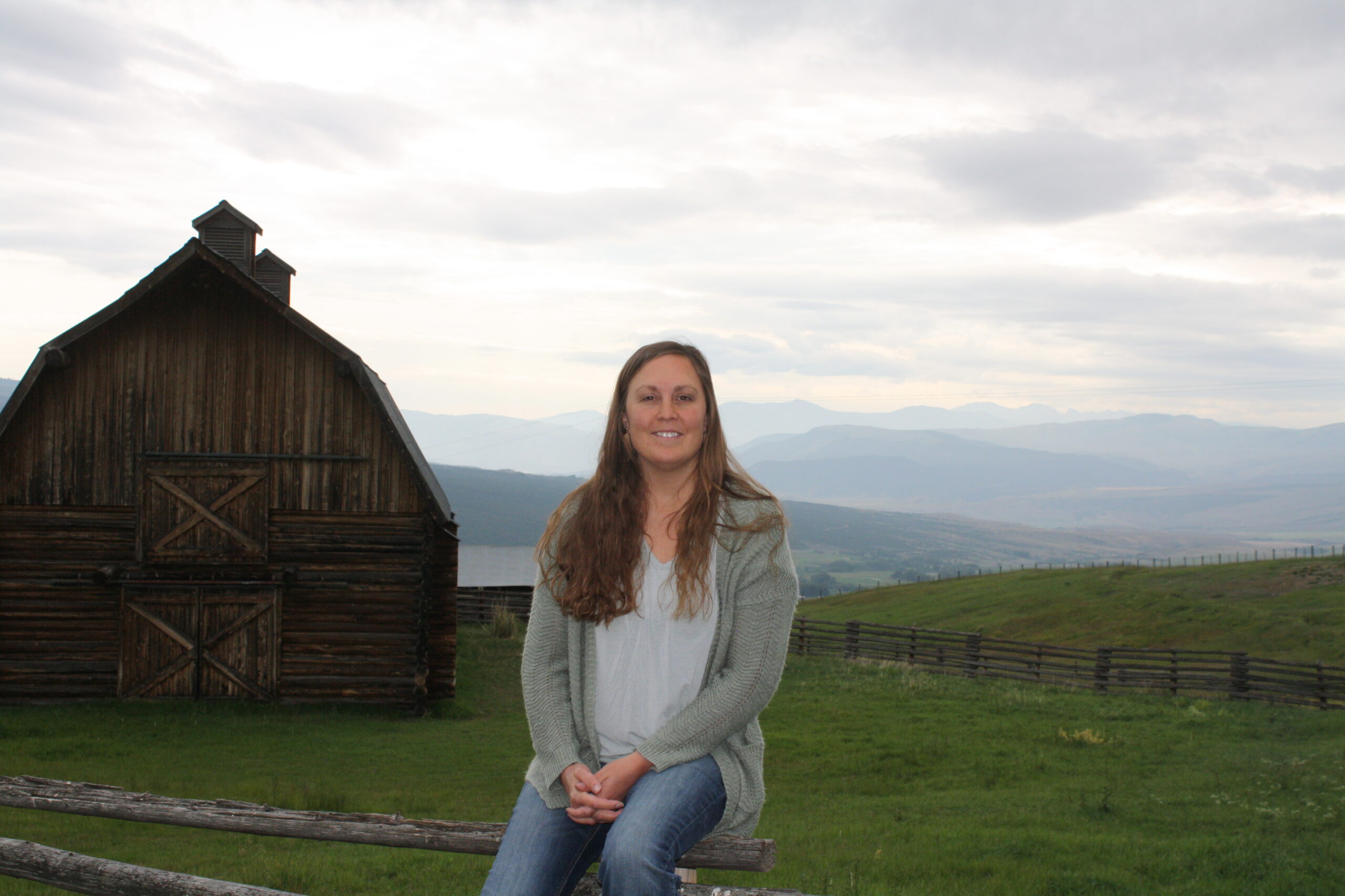 Picture is of a lady sitting on a fence with a barn and mountains behind her.