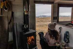A tiny woodstove is near the door in an RV. A lady and her dog are sitting at the open door looking towards snow covered mountains.