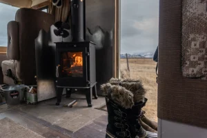A DIY Tiny Wood Stove is featured near a door of a motorhome. The door is open and overlooking mountains. A pair of fluffy winter boots are near the door.