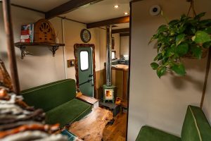 A green woodstove is near the entry door within a camper. Green couches and a hanging plant add to the atmosphere.